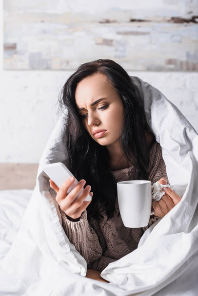 beteg fiatal barna nő forró ital és szövet segítségével smartphone az ágyban - Fotó, kép