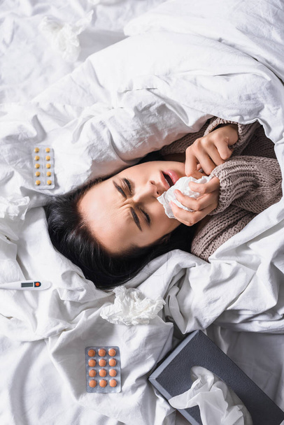 vista superior de la mujer morena joven enferma con tejido, pastillas y termómetro en la cama - Foto, Imagen