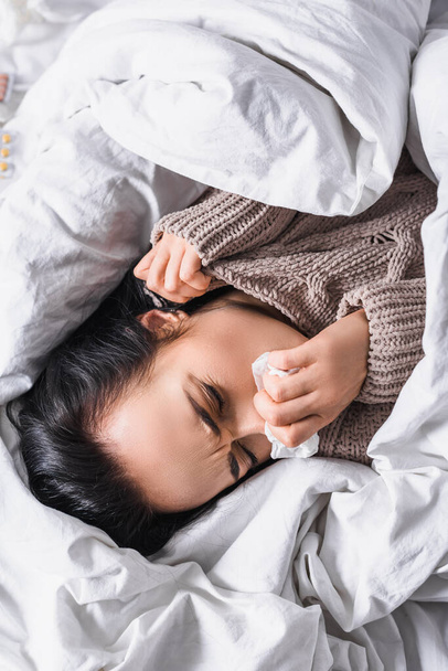 πάνω όψη της άρρωστης νεαρής μελαχρινής γυναίκας με ιστό στο κρεβάτι - Φωτογραφία, εικόνα