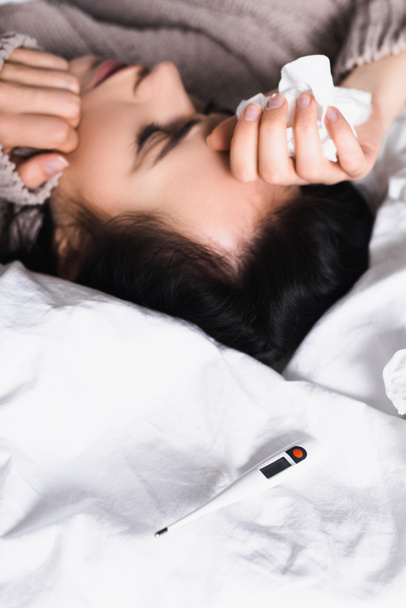 άρρωστη νεαρή μελαχρινή γυναίκα με θερμόμετρο και ιστό στο κρεβάτι - Φωτογραφία, εικόνα