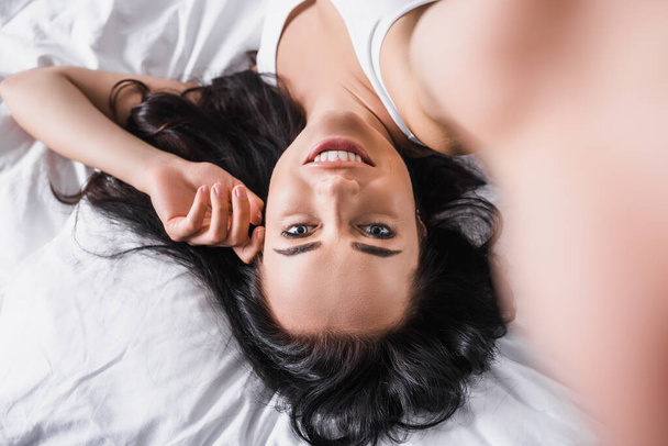 top view of young smiling brunette woman lying in bed - Foto, afbeelding