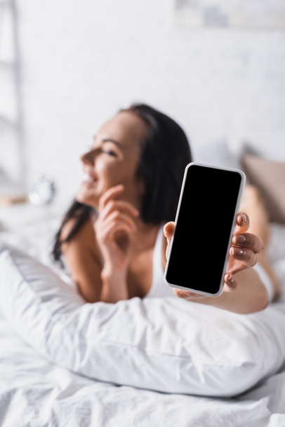 young happy brunette woman in bed presenting smartphone, blurred background - Φωτογραφία, εικόνα