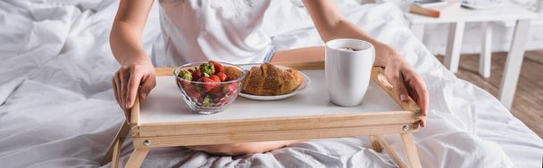 ベッドで朝食のためにクロワッサンとイチゴを持っている若い女性のクロップドビュー、バナー - 写真・画像