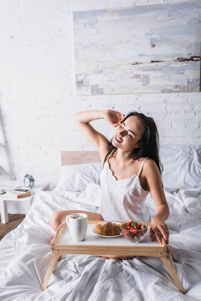 young brunette woman having croissant and strawberry for breakfast in bed - Photo, Image
