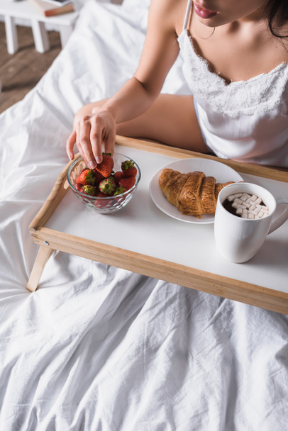 cropped view of young brunette woman having cocoa, croissant and strawberry for breakfast in bed - Photo, Image
