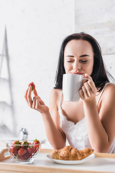 Genç esmer kadın kahvaltıda kruvasan ve çilek yiyor ve yatakta kakao içiyor. - Fotoğraf, Görsel
