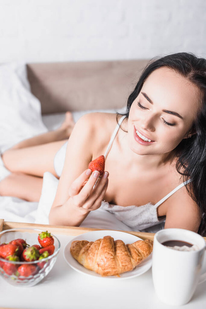 junge brünette Frau mit Croissant, Erdbeere und Kakao zum Frühstück im Bett - Foto, Bild