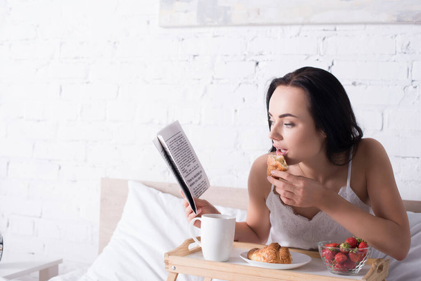 junge brünette Frau frühstückt mit Croissant, Erdbeere und Kakao, während sie im Bett Zeitung liest - Foto, Bild