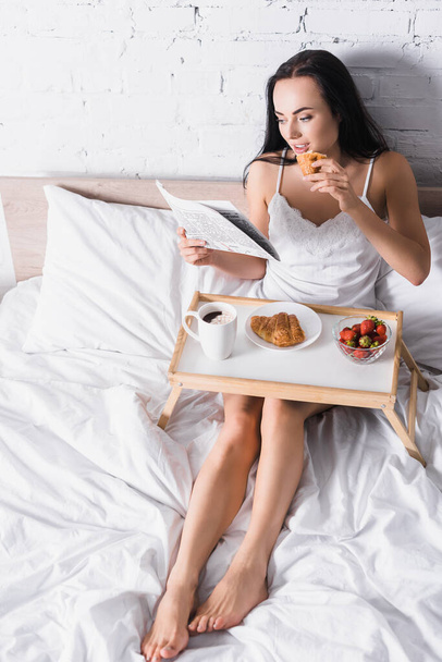 young brunette woman having croissant, strawberry and cocoa for breakfast while reading newspaper in bed - Foto, Bild