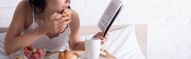 περικοπή άποψη της μελαχρινή γυναίκα έχοντας κρουασάν, φράουλα και κακάο για πρωινό, ενώ διαβάζετε εφημερίδα στο κρεβάτι, πανό - Φωτογραφία, εικόνα