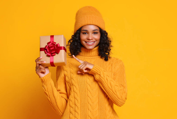 Mujer étnica alegre en suéter de punto y sombrero sonriendo y mirando a la cámara mientras señala regalo envuelto durante la celebración de vacaciones contra fondo amarillo - Foto, Imagen