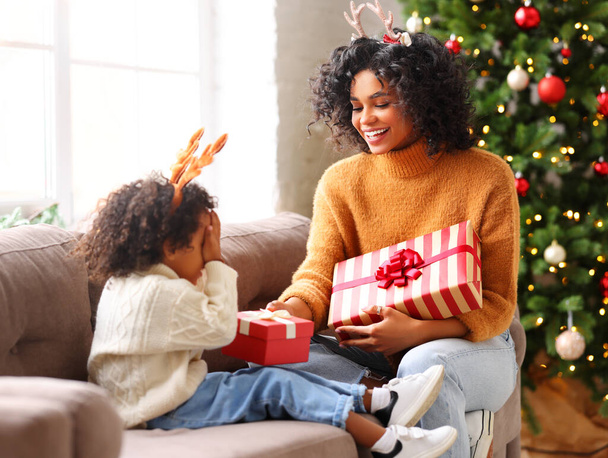 Happy ethnic woman with deer antlers giving present to cute boy covering eyes while sitting on couch on Christmas da - Φωτογραφία, εικόνα
