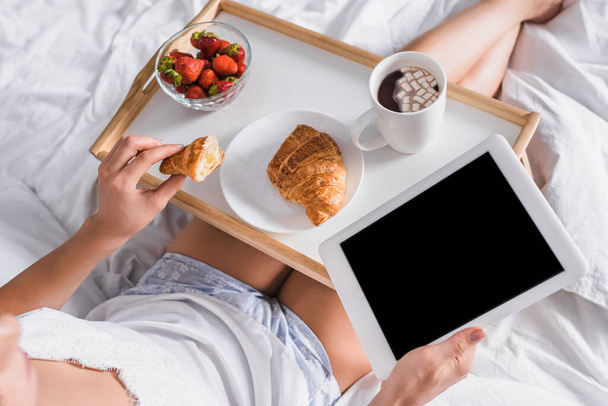 przycięty widok kobiety z rogalikiem, truskawką i kakao na śniadanie trzymając cyfrową tabletkę w łóżku - Zdjęcie, obraz