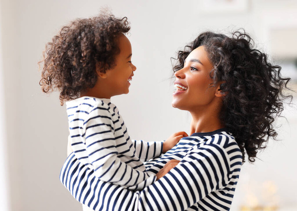 Side view of delighted ethnic woman smiling and carrying cheerful boy with curly hair while spending time at home togethe - 写真・画像