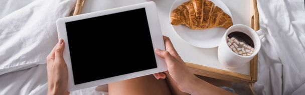 cropped view of woman having croissant and cocoa for breakfast while holding digital tablet in bed, banner - Foto, imagen