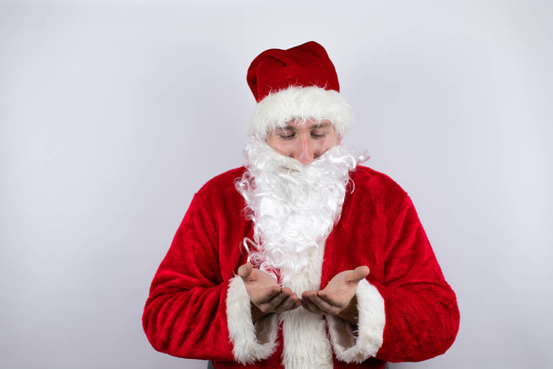 Als Weihnachtsmann verkleideter Mann, der vor isoliertem weißem Hintergrund steht, lächelnd die Hände in den Händen hält und Gesten entgegennimmt oder gibt. Halt und Schutz - Foto, Bild