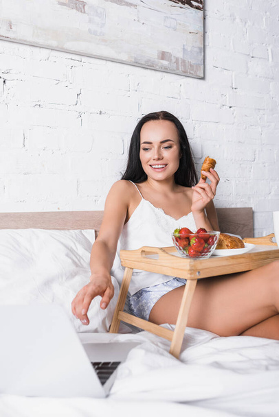 smiling young brunette woman having breakfast in bed while using laptop - Φωτογραφία, εικόνα
