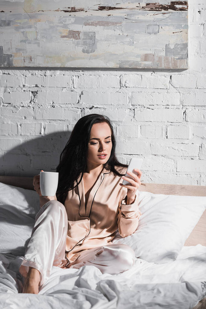 Genç esmer kadın sabahları elinde kupa ve akıllı telefonuyla yatakta oturuyor. - Fotoğraf, Görsel