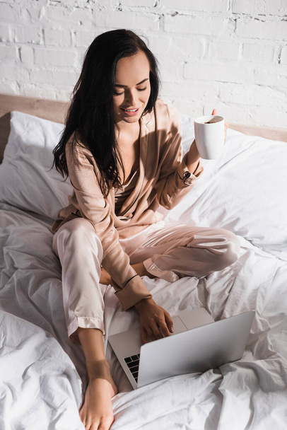 young brunette woman sitting in bed with mug and laptop at morning - Fotó, kép