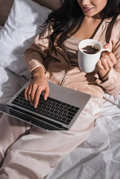 若いブルネットの女性が朝にマグカップとラップトップでベッドに座っているのをクロップドビュー - 写真・画像