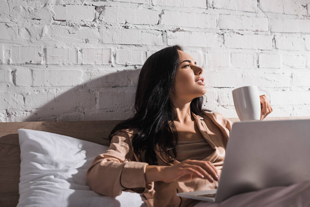 mladá brunetka žena sedí v posteli s hrnkem a notebookem v dopoledních hodinách - Fotografie, Obrázek