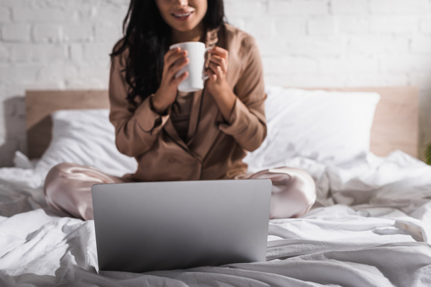 Genç esmer bir kadının sabahları kupa ve dizüstü bilgisayarla yatakta oturduğu görüntüler. - Fotoğraf, Görsel