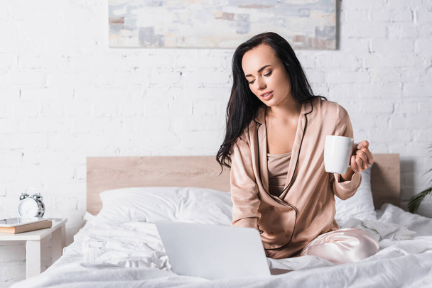 young brunette woman sitting in bed with mug and laptop at morning - Φωτογραφία, εικόνα