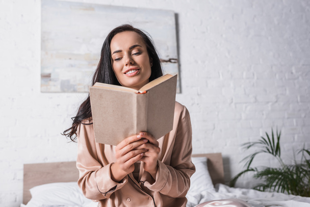 smiling young brunette woman sitting in bed with book at morning - Foto, imagen