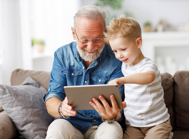 Szczęśliwa rodzina starszy dziadek i chłopiec siedzi na kanapie i oglądanie kreskówek na tablecie w weekend w domu razem - Zdjęcie, obraz