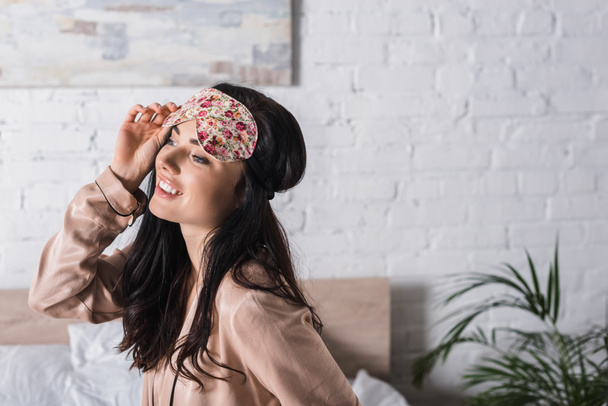 smiling young brunette woman sitting in bed in sleep mask - Foto, afbeelding