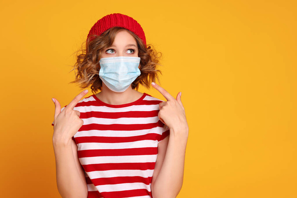 Joven hembra en traje de moda apunta a la máscara médica mirando el espacio vacío contra el fondo amarillo durante pandemi - Foto, imagen