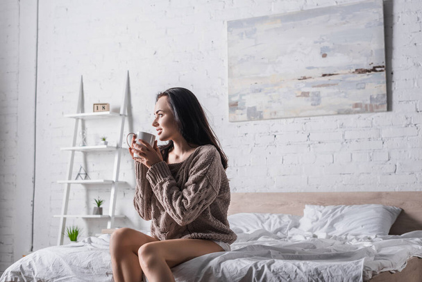 mladá brunetka žena ve svetru sedí v posteli s hrnkem horkého kakaa ráno - Fotografie, Obrázek