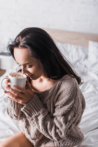 young brunette woman in sweater sitting in bed with mug of hot cocoa at morning - Foto, afbeelding