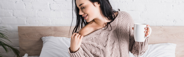 young brunette woman in sweater with bare shoulder sitting in bed with mug of hot cocoa at morning, banner - Фото, зображення