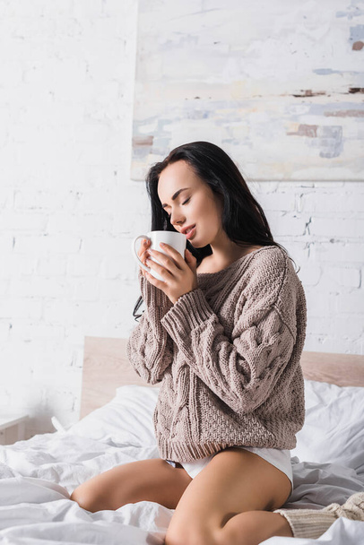 sexy young brunette woman in sweater sitting in bed with mug of hot cocoa at morning - Φωτογραφία, εικόνα