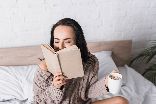 happy young brunette woman in sweater sitting in bed with mug of hot cocoa and book at morning - Фото, зображення