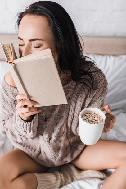 young brunette woman in sweater sitting in bed with mug of hot cocoa and book at morning - Foto, imagen