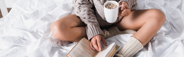 vista recortada de la mujer en suéter y calcetines de punto sentado en la cama con taza de cacao caliente y libro por la mañana, pancarta - Foto, Imagen