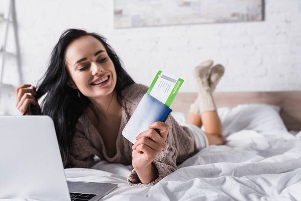 heureuse jeune femme brune en pull couché dans le lit avec ordinateur portable et billet d'avion - Photo, image