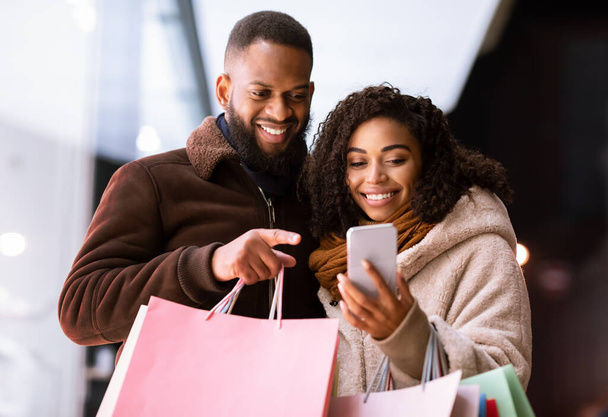 Retrato de feliz pareja negra usando teléfono con bolsas de compras - Foto, imagen