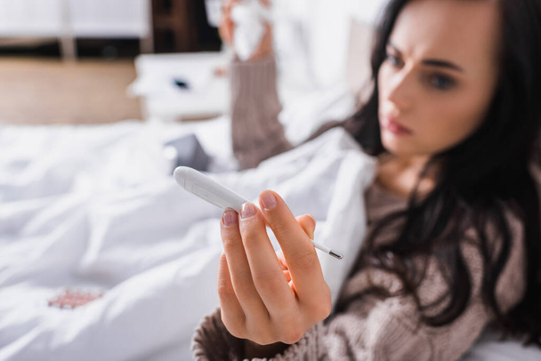 blurred ill young brunette woman in sweater lying in bed with thermometer and tissue - Photo, Image