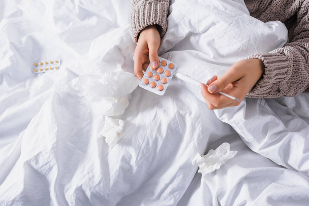 vista cortada de mulher jovem doente com tecido, pílulas e termômetro na cama - Foto, Imagem