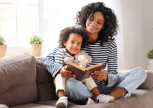 Happy ethnic family: delighted african american mother sitting on couch and reading interesting book to cute  son on weekend day at hom - Фото, изображение