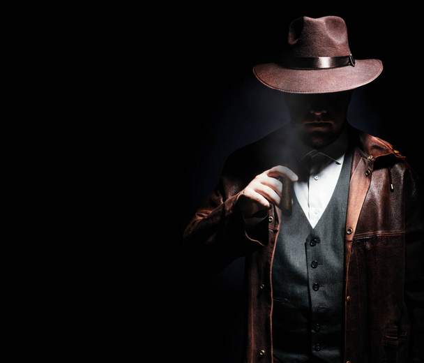 Foto de un detective sombreado con chaqueta y sombrero sosteniendo y fumando cigarro sobre fondo negro. - Foto, Imagen