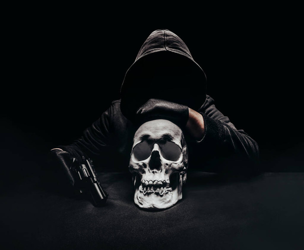 Photo of a creepy horror dark killer with revolver gun sitting with human skull on a table. - Фото, изображение