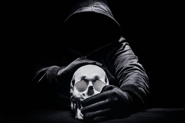 Horror-Foto eines beängstigenden Mannes in Kapuzenpulli, der sitzt und den menschlichen Schädel zum Schweigen bringt. - Foto, Bild
