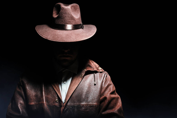 Φωτογραφία ενός σκιασμένου ντετέκτιβ με σακάκι και καπέλο σε μαύρο φόντο. - Φωτογραφία, εικόνα