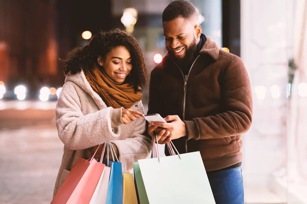 Retrato de feliz pareja negra usando smartphone sosteniendo bolsas de compras - Foto, Imagen