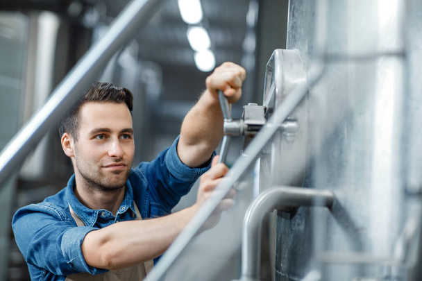 Millennial attractive male worker or owner in apron turns valve on big boiler for beer in interior of plant - Photo, Image