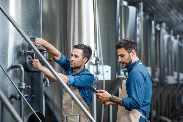 Millennial guy worker in apron turns a valve on big boiler for lager, manager with tablet controls process in interior - Photo, Image